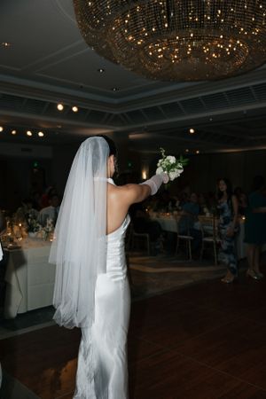 A bride in a white strapless gown and long veil holds up her bouquet under an elegant chandelier in a ballroom.