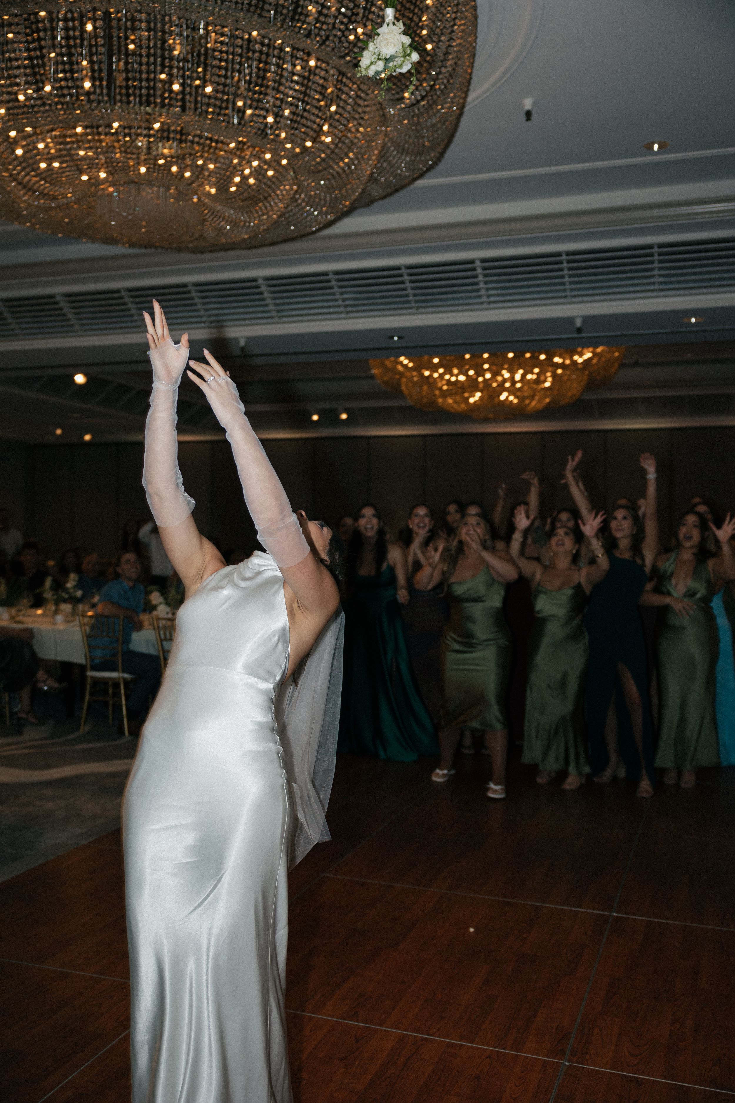 A wedding reception with a person in a white gown dancing beneath an ornate chandelier.