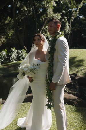 Newlywed couple wearing white poses outdoors in garden setting with flowing veil and traditional Hawaiian lei decorations.