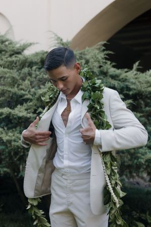 Groom in an elegant white suit and floral lei adjusts his jacket while standing in a garden setting.