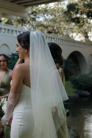 A bride in a fitted white dress and long flowing veil stands by a reflection pond with arched architecture in the background.
