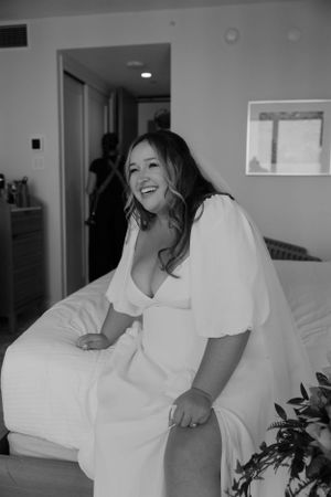 A joyful bride in a white wedding dress sitting on a bed during wedding preparations.