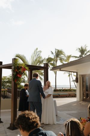 Outdoor wedding ceremony under wooden pergola with tropical palm trees and ocean backdrop.