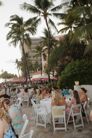 Wedding reception setup with white chairs under palm trees at a beachfront venue.