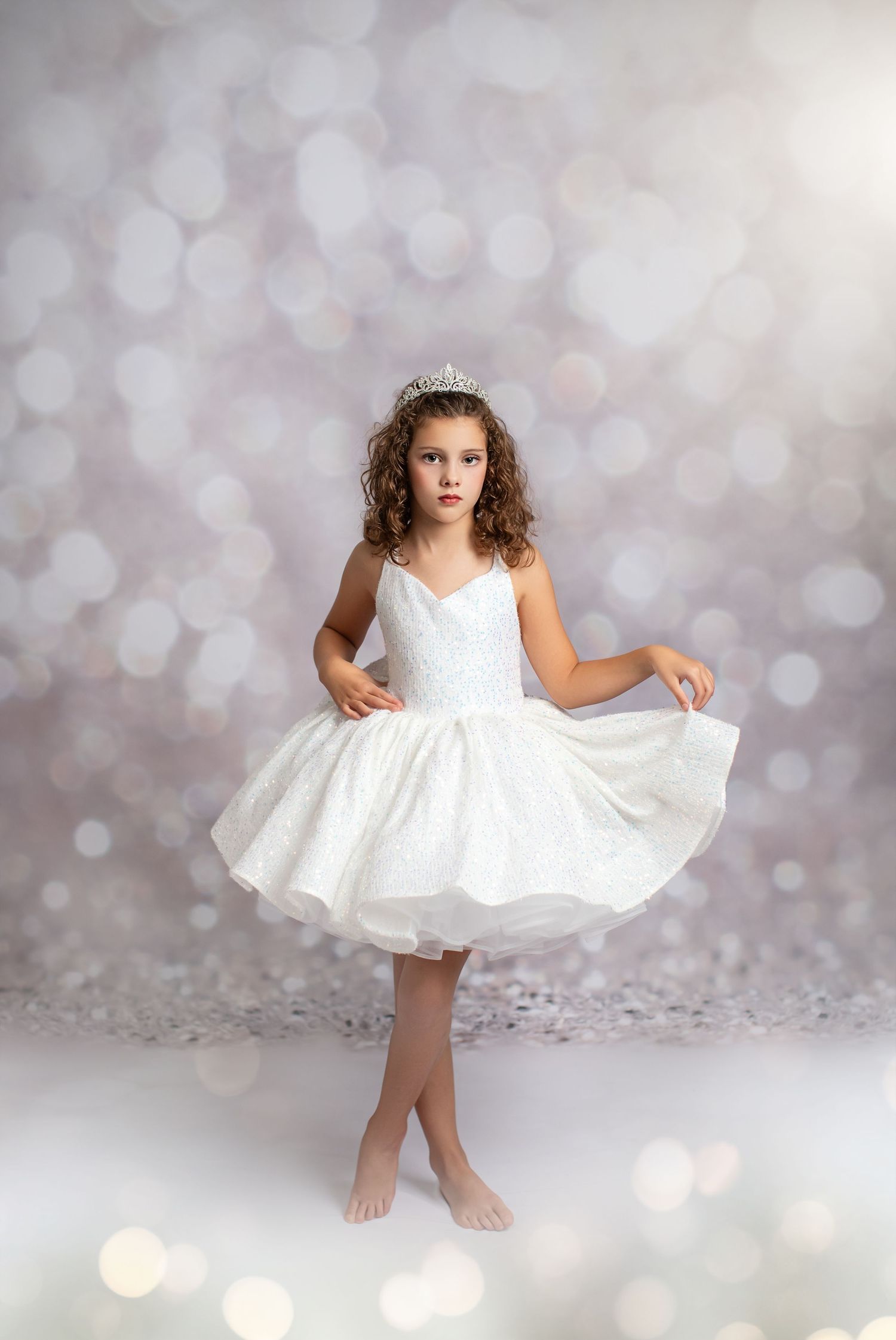 Young dancer in a white tulle dress and tiara poses gracefully barefoot against a bokeh backdrop.