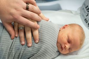 Gentle hands touching sleeping newborn wrapped in soft gray blanket.