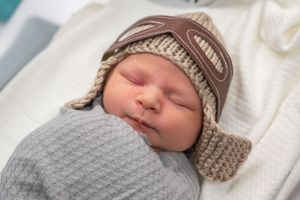 Peaceful sleeping newborn wearing brown knit hat against white bedding.