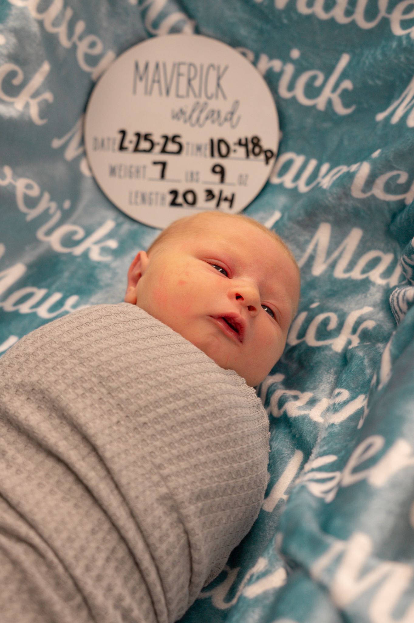 Newborn wearing a gray blanket resting on teal patterned hospital bedding.