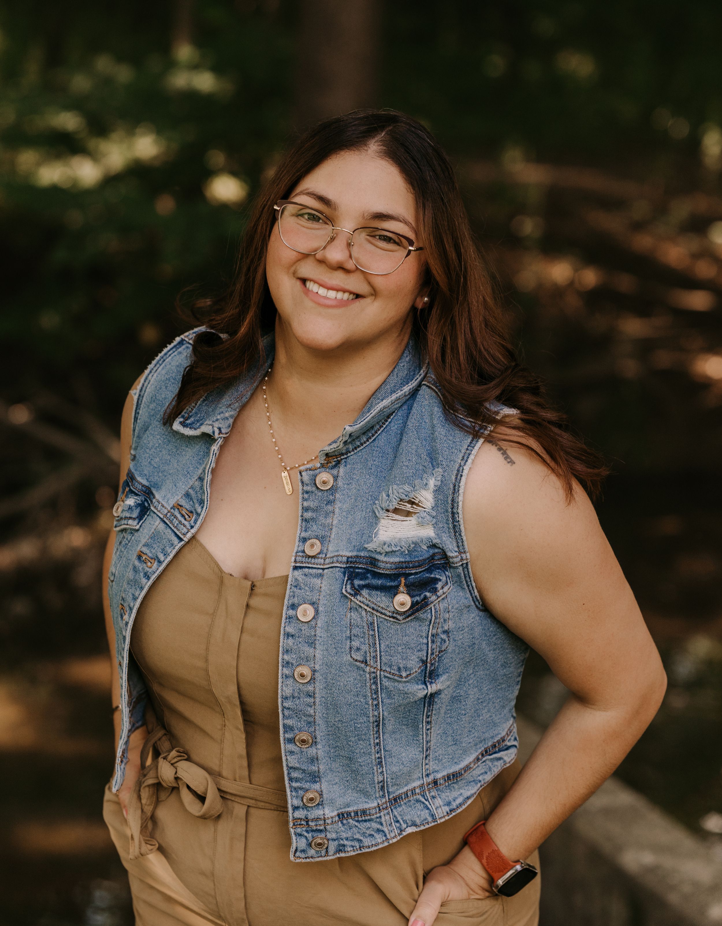 Portrait of person in denim vest and tan top smiling outdoors.