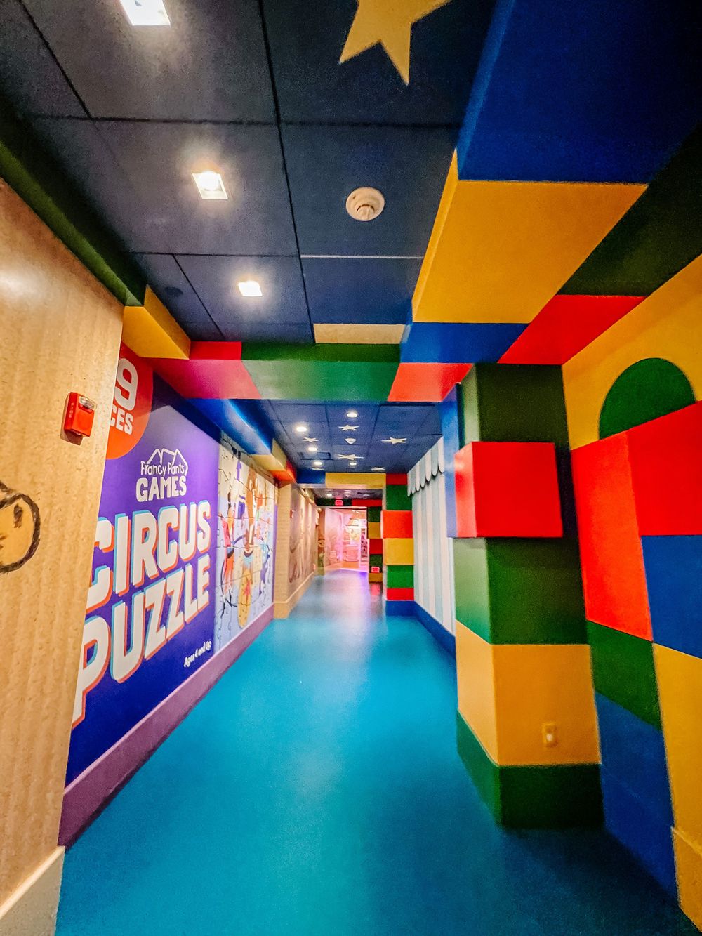 Colorful hallway decorated with bright geometric shapes and circus puzzle graphics on walls with a blue floor and star accent.
