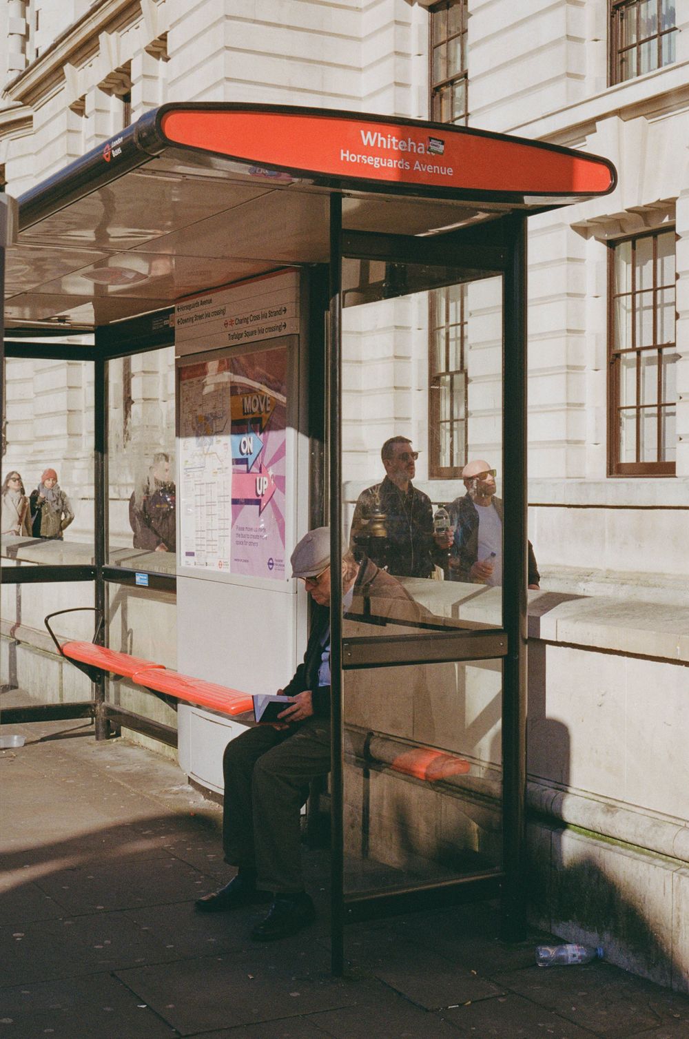 A red and black bus shelter with posted notices stands outside a classical building.