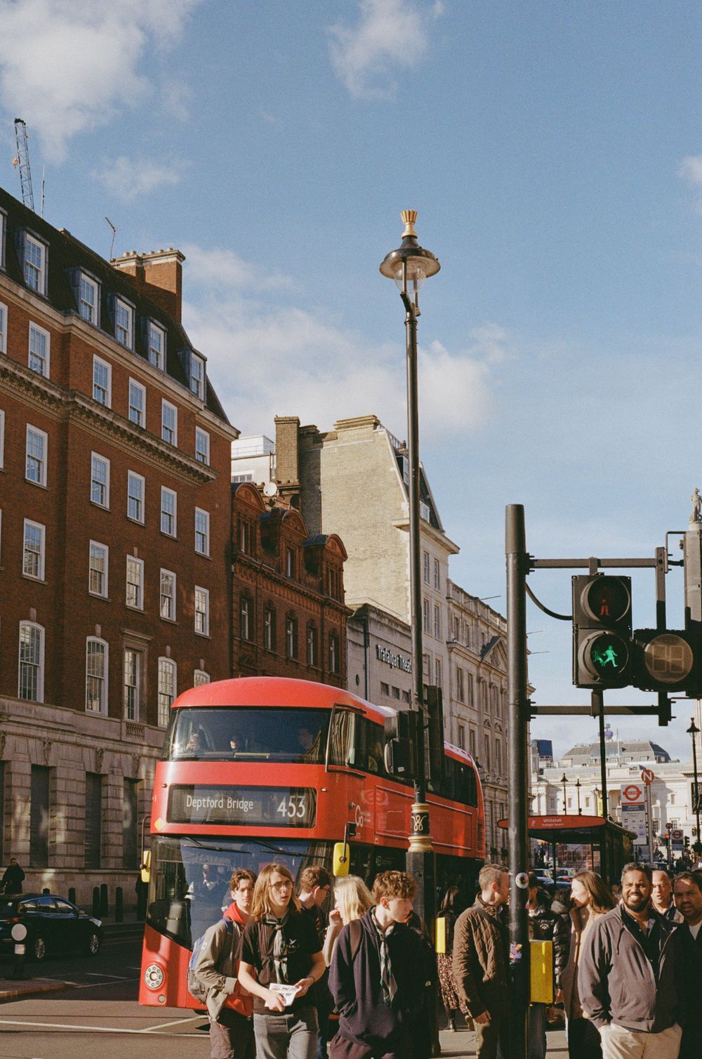 A red double-decker bus stops at a busy London street corner with pedestrians waiting to cross.