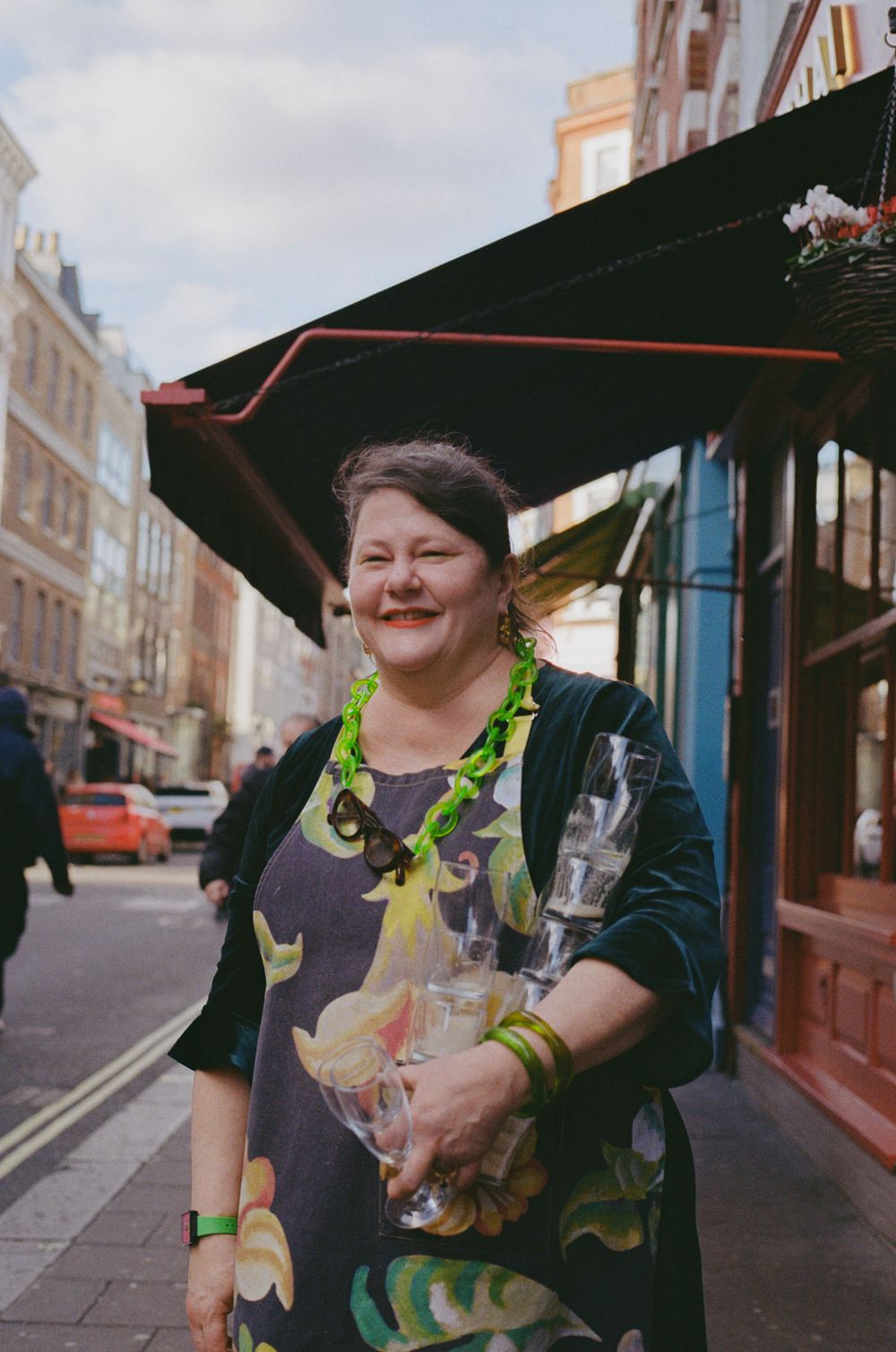 A person in colorful attire holds a bottle while standing under a black awning on a city street.