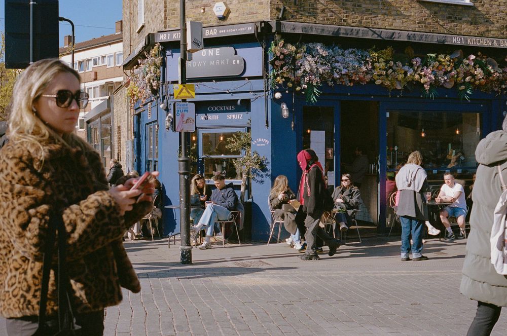 Urban street corner with shoppers passing by storefronts on a sunny day.