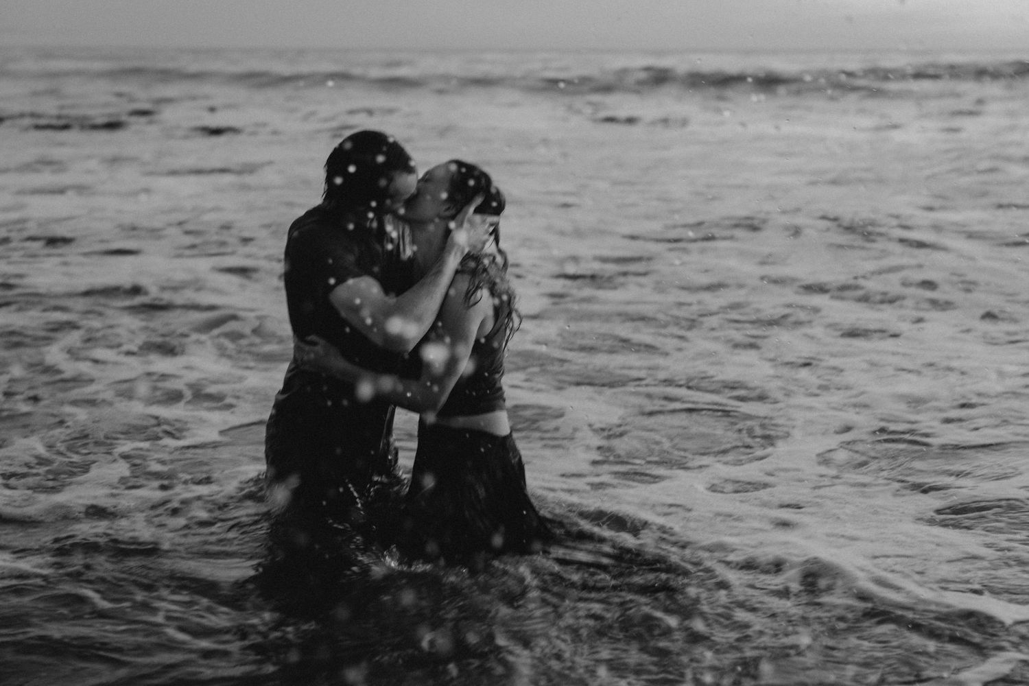 A romantic black and white photograph shows a couple embracing while standing in ocean waves.
