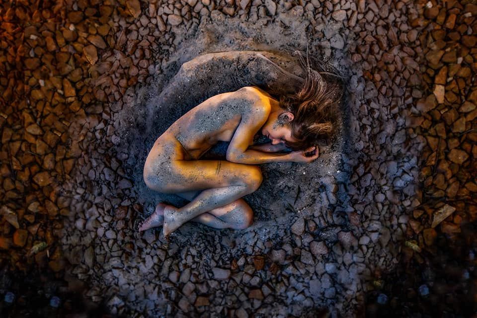 Artistic nude figure curled in fetal position surrounded by dark textured pattern in overhead view.