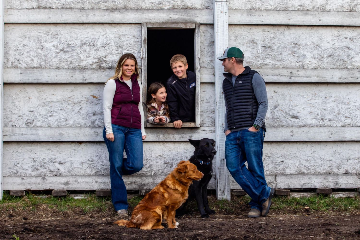 Family standing by a rustic white barn wall with a brown dog and small pig in a window.
