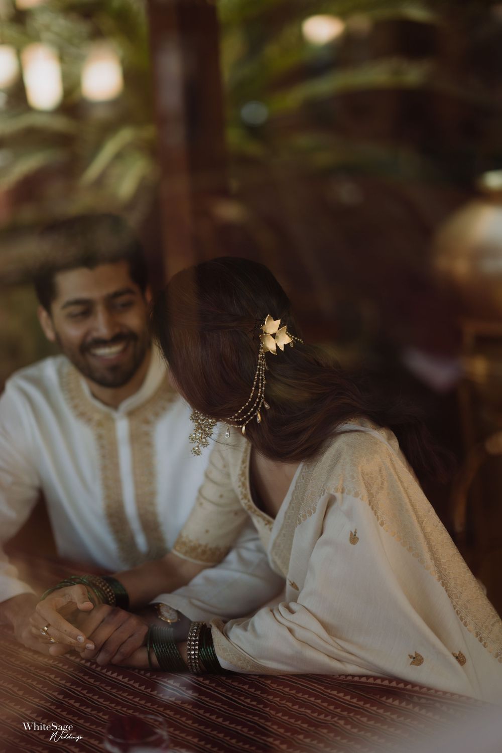 Couple in white ethnic wear sharing a joyful moment, bride adorned with gold hair accessories at their wedding.