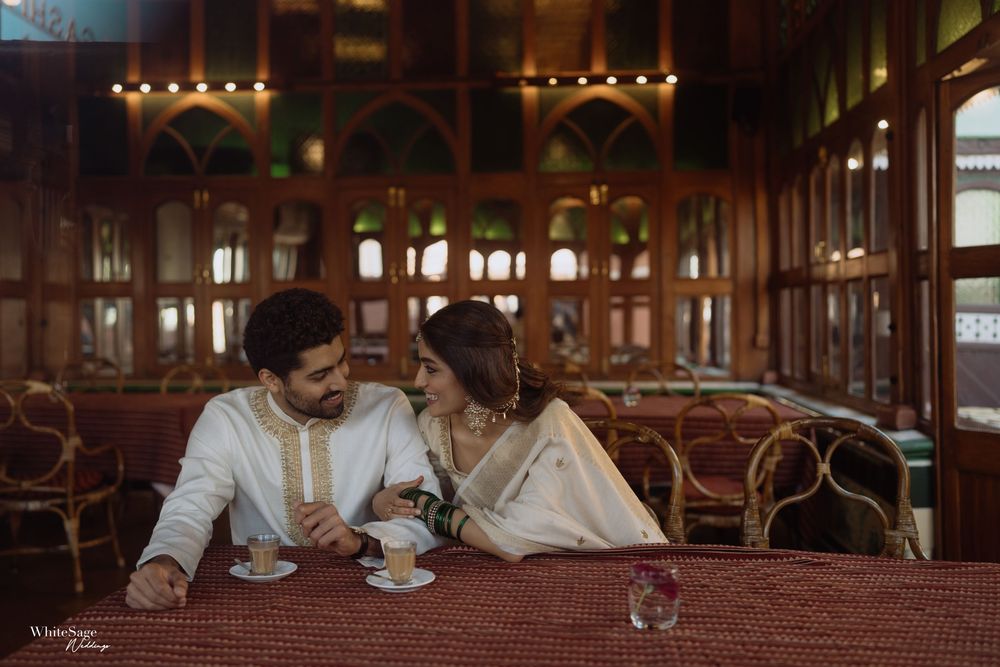 Couple sharing an intimate moment at a vintage wooden cafe table, dressed in elegant white traditional attire.