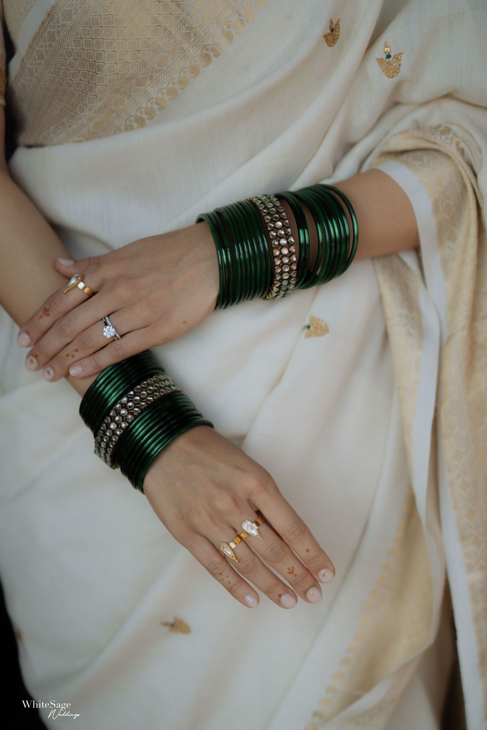 Bride wearing dark green bangles with rhinestones and gold rings, draped in an ivory silk saree with gold motifs.