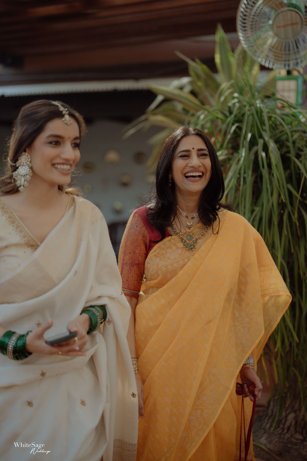 Two women in elegant sarees, one ivory and one yellow, smiling joyfully at an Indian wedding celebration.