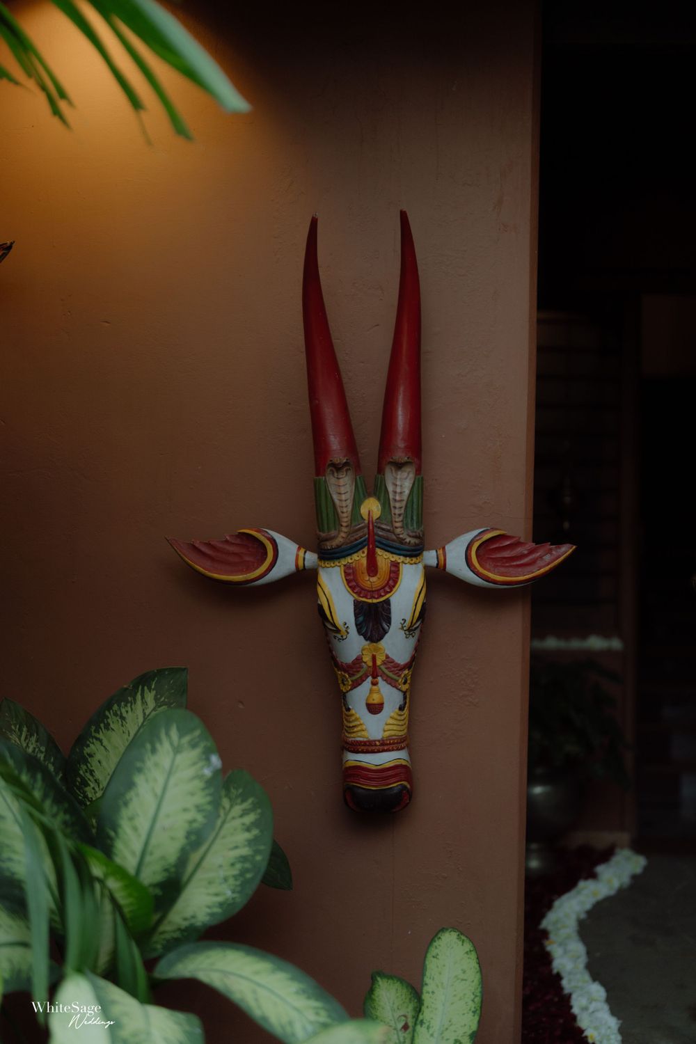 A colorful tribal mask decoration mounted on a brown wall with plants in the foreground.