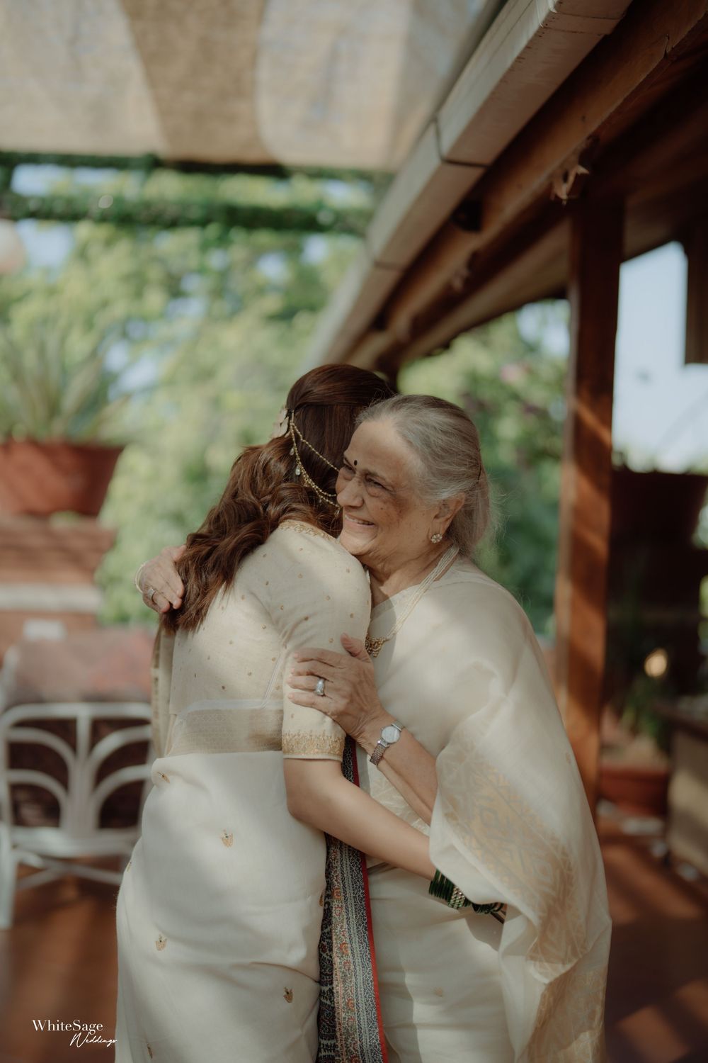 Two women in elegant cream sarees share a warm embrace at an outdoor wedding venue surrounded by lush greenery.