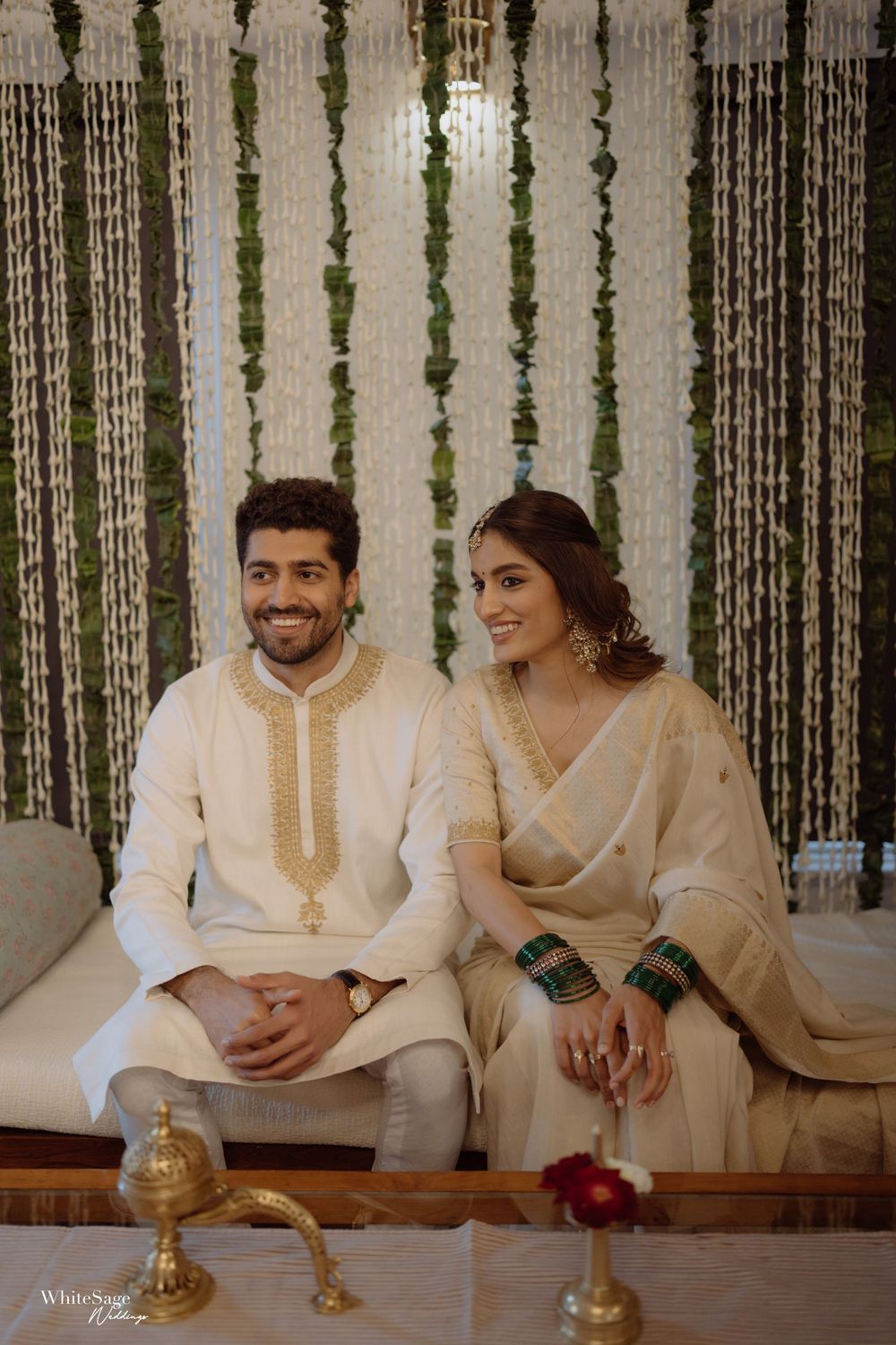 Indian couple in matching cream and gold outfits seated together at a traditional engagement ceremony with floral backdrop.