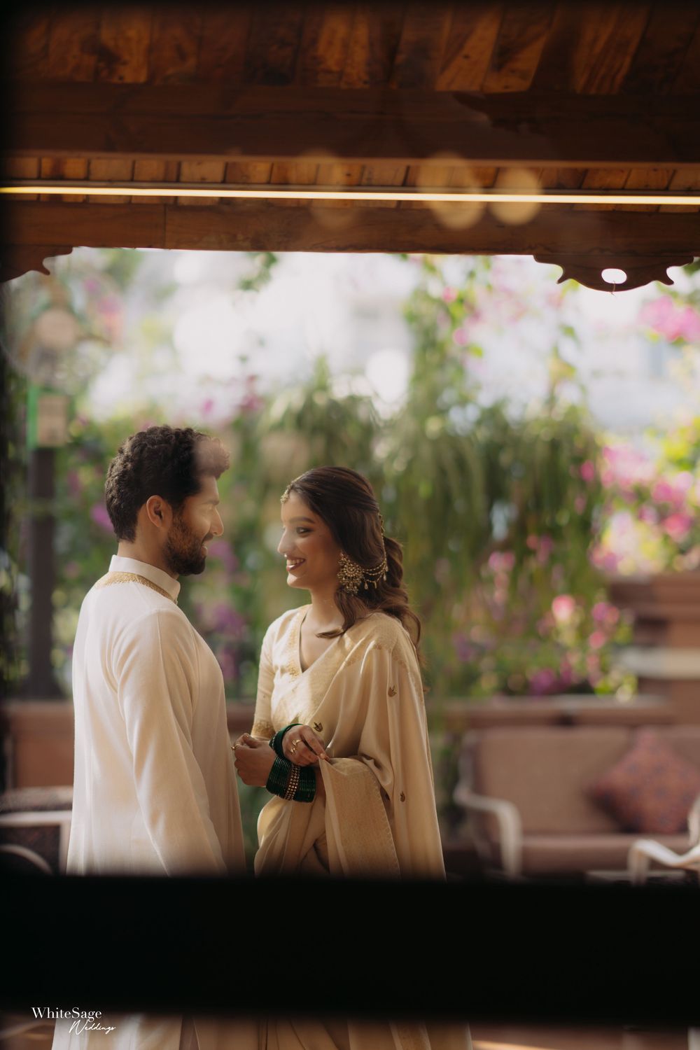 Couple in cream traditional Indian attire sharing a romantic moment in a lush garden setting.