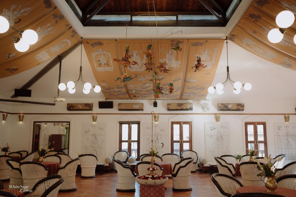Elegant colonial-style hall with ornate painted ceiling mural, rattan furniture, chandeliers, and large windows.