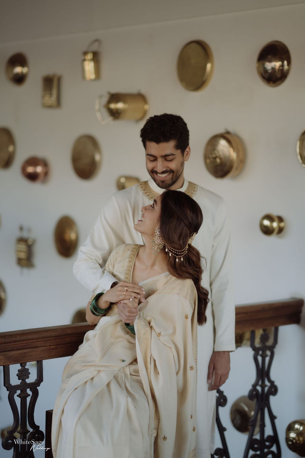 Couple in cream traditional Indian attire posing romantically against a white wall decorated with brass plates.