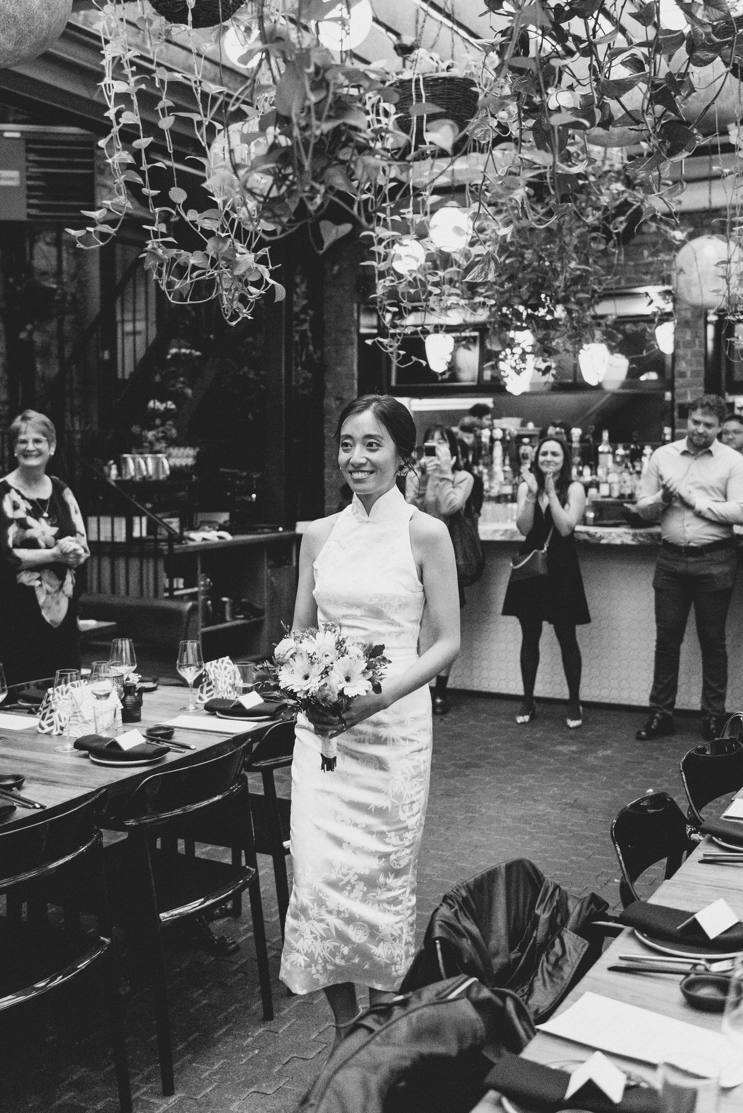 Beautiful bride in traditional qipao dress holds bouquet at wedding reception with hanging lights and decorations overhead.