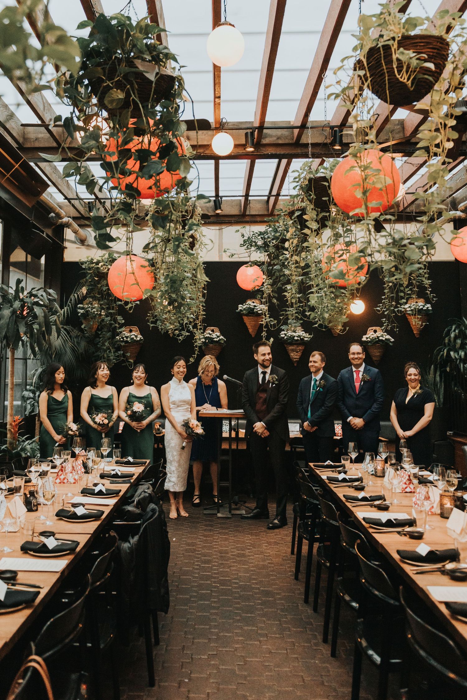 Elegant wedding reception setup in rustic greenhouse venue with hanging paper lanterns and lush greenery decorations.