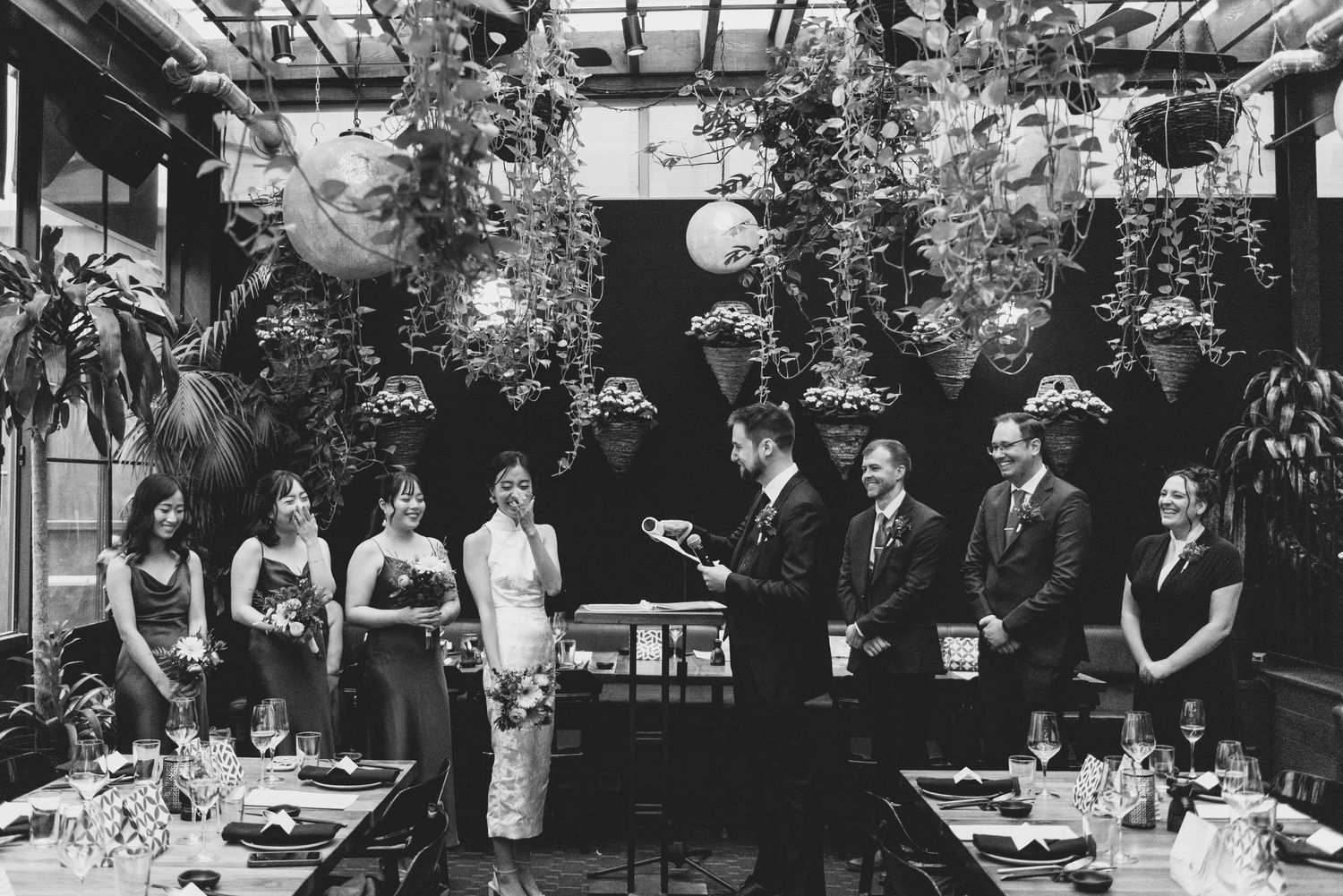 A black and white photograph of a formal dining event with several people gathered in a plant-filled room with lanterns.