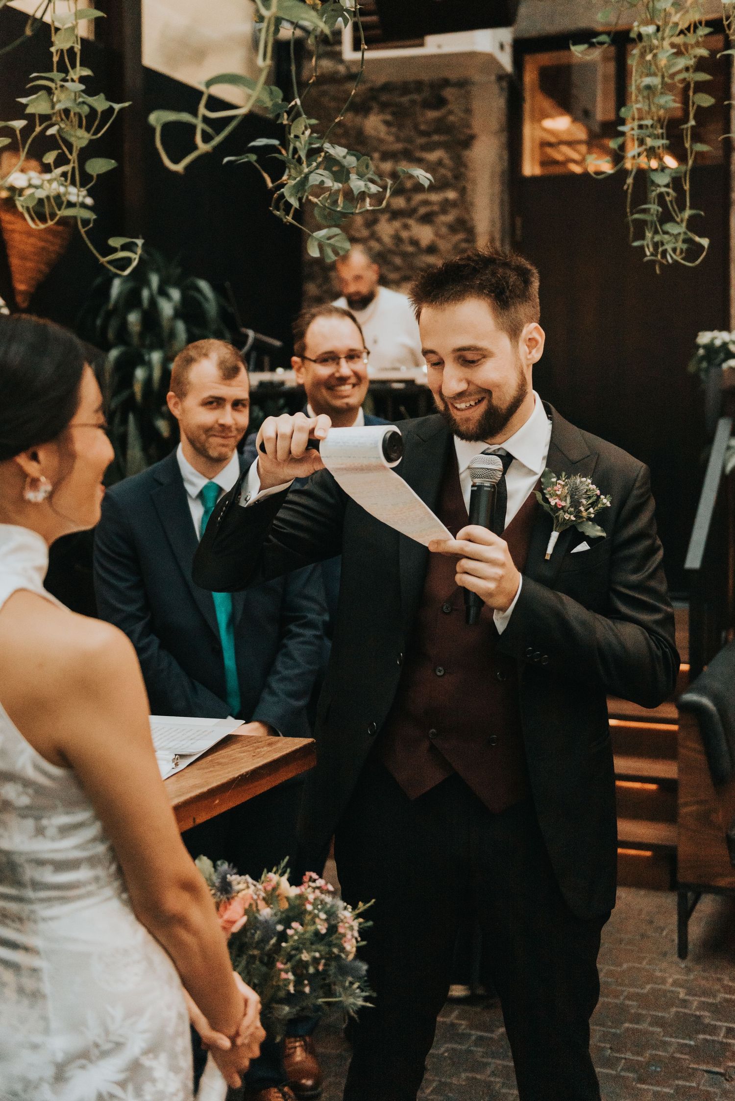 At an intimate outdoor wedding ceremony, vows are exchanged surrounded by hanging plants and decorative lighting.