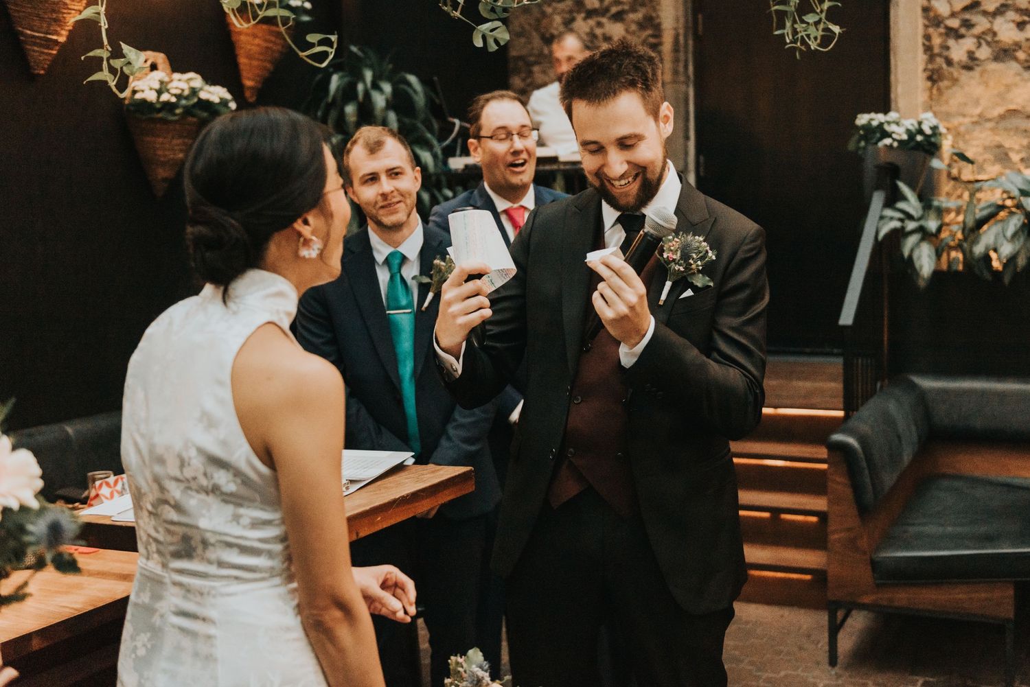 A couple shares a laugh during a joyful wedding reception moment while guests smile in the background.