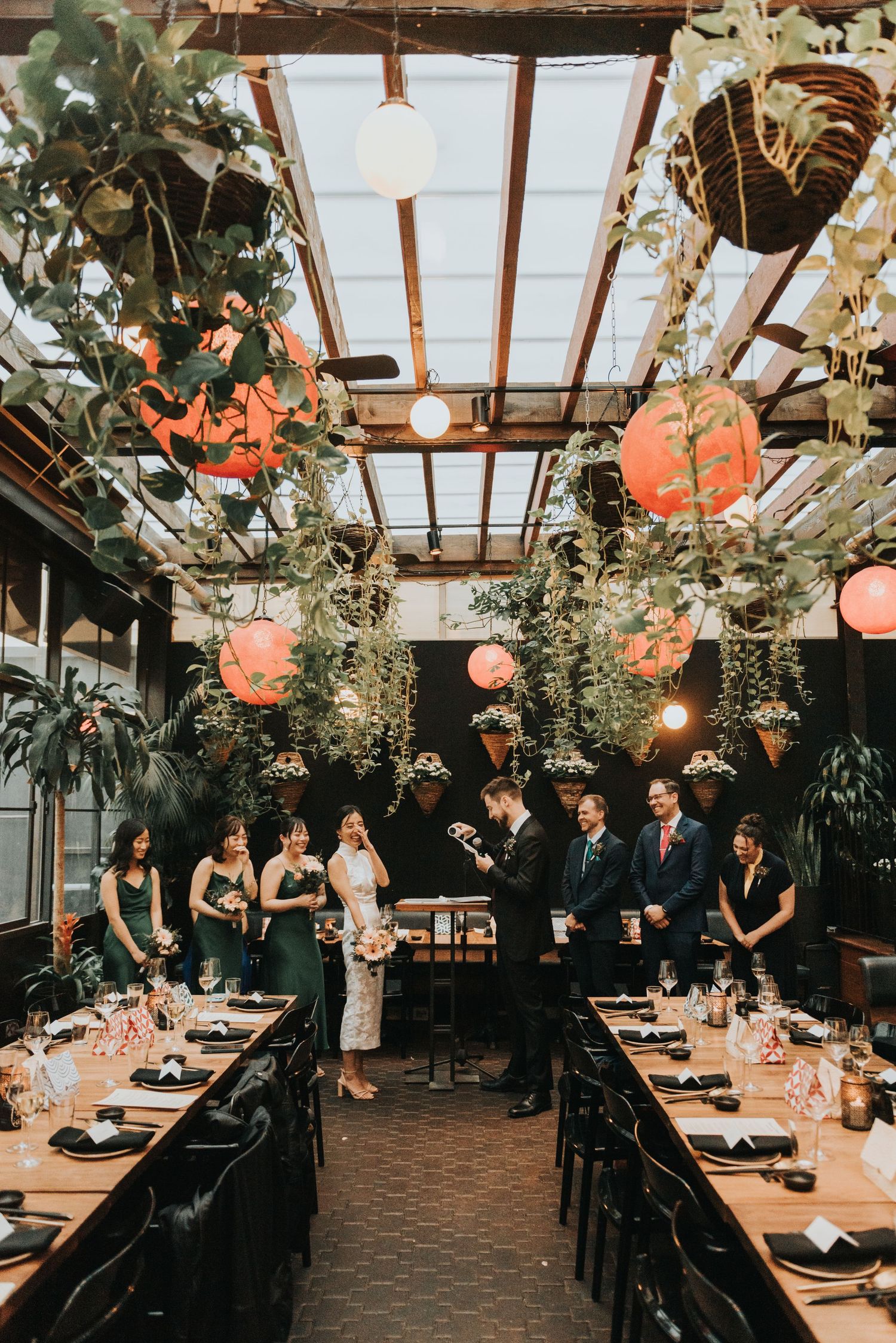 Wedding reception in a greenhouse-style venue with hanging plants, paper lanterns, and rustic wooden beams overhead.