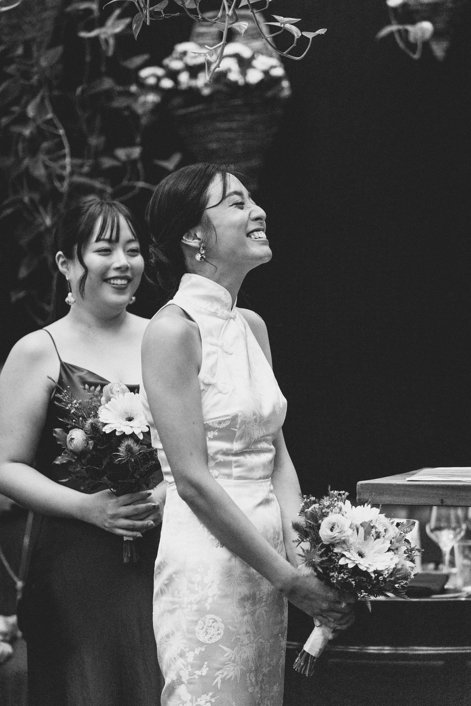 Black and white photo of joyful wedding celebration with two people in formal attire holding bouquets and sharing laughter.