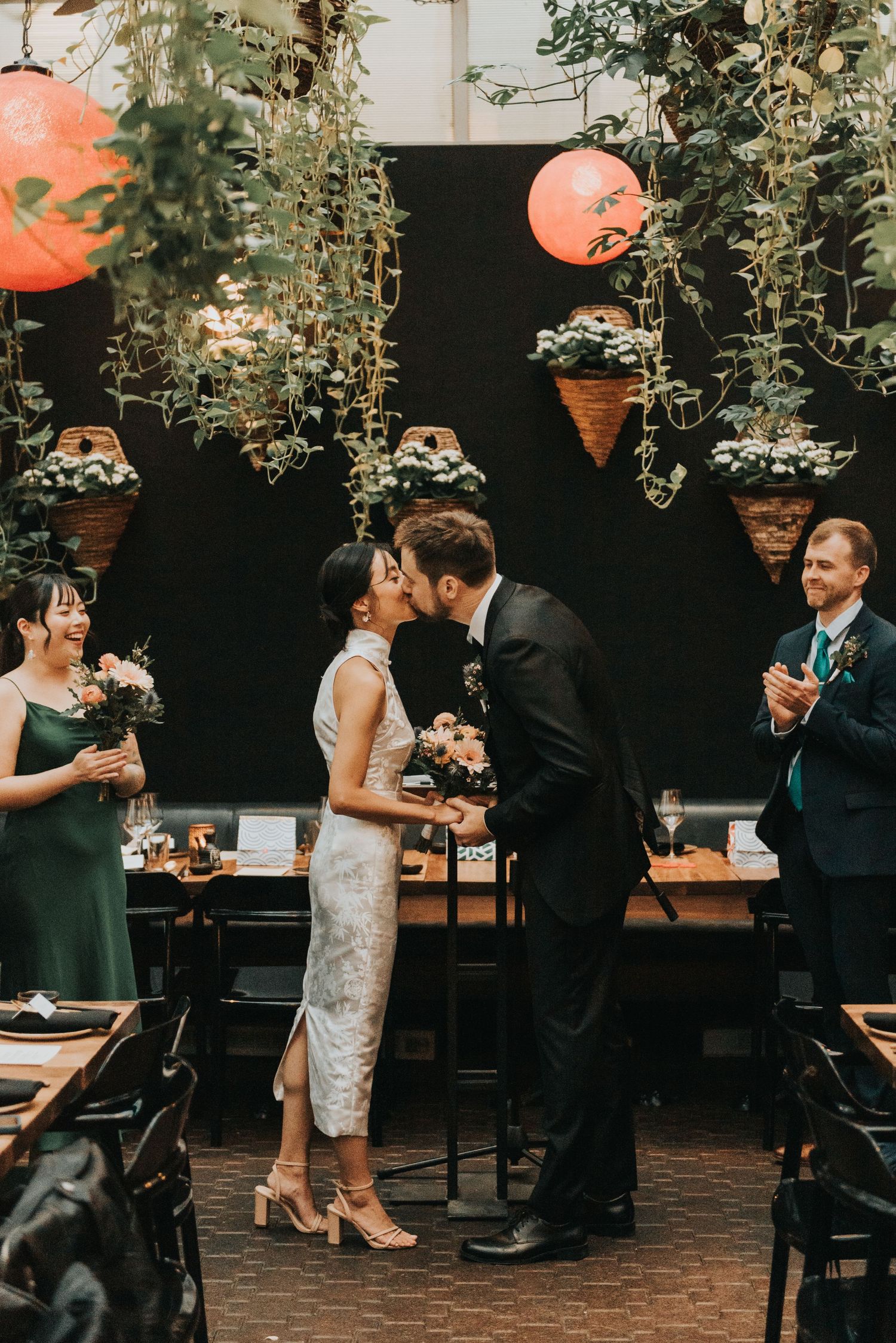 Intimate wedding ceremony in a garden-themed restaurant with hanging plants and orange paper lanterns against a dark backdrop.