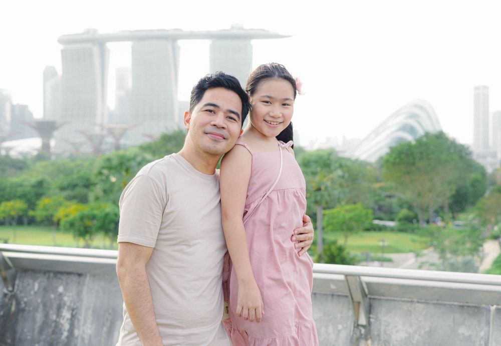 Two people pose together outdoors with Marina Bay Sands and Singapore city skyline visible in the background.