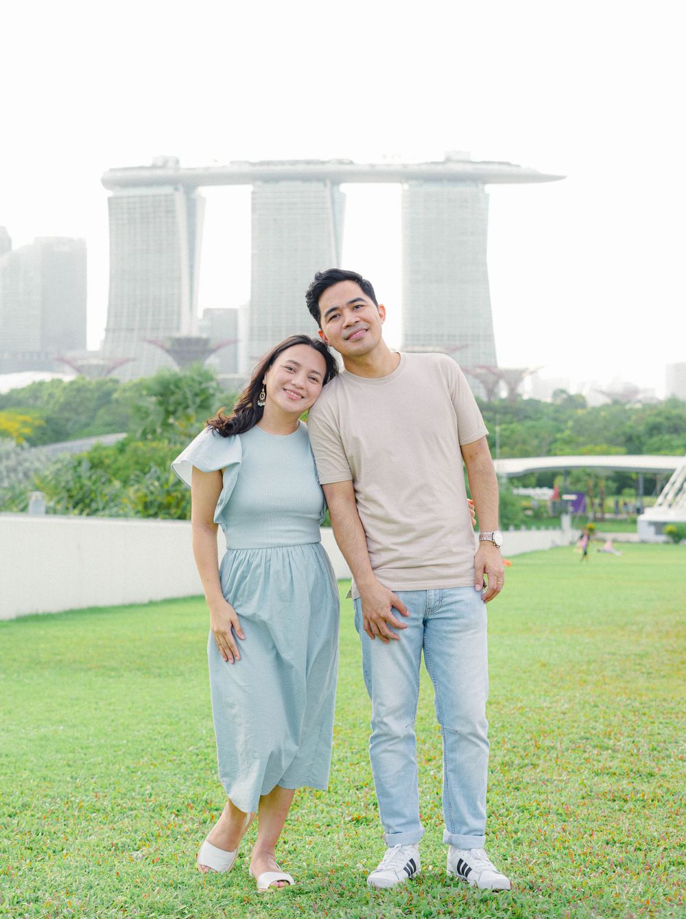 A couple stands together on grass with Singapore's Marina Bay Sands hotel towers visible in the background.