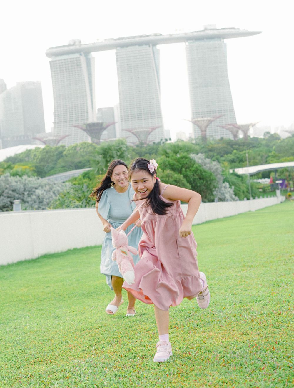 Two friends in flowy dresses playfully run and pose on a grassy hill with Singapore's Marina Bay Sands visible in the background.