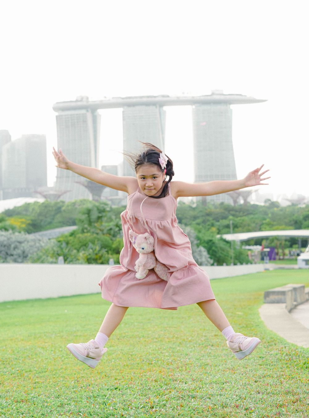 A child in a pink dress jumps joyfully on grass with Marina Bay Sands hotel visible in the background in Singapore.