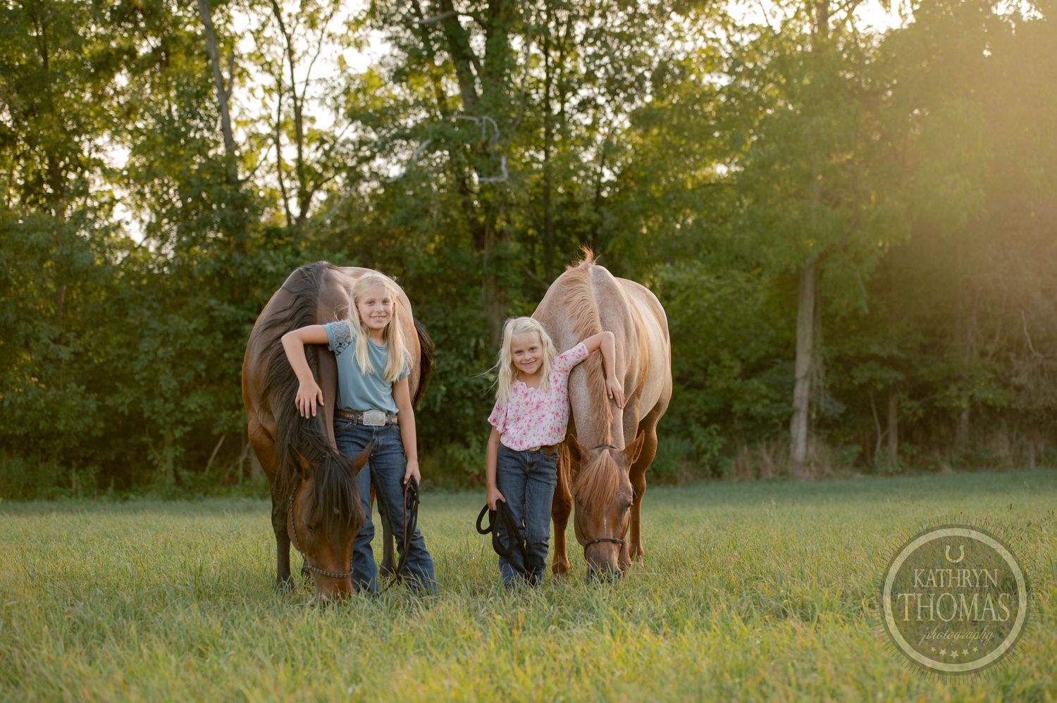 Two people stand with their horses as they graze in a sunlit field.