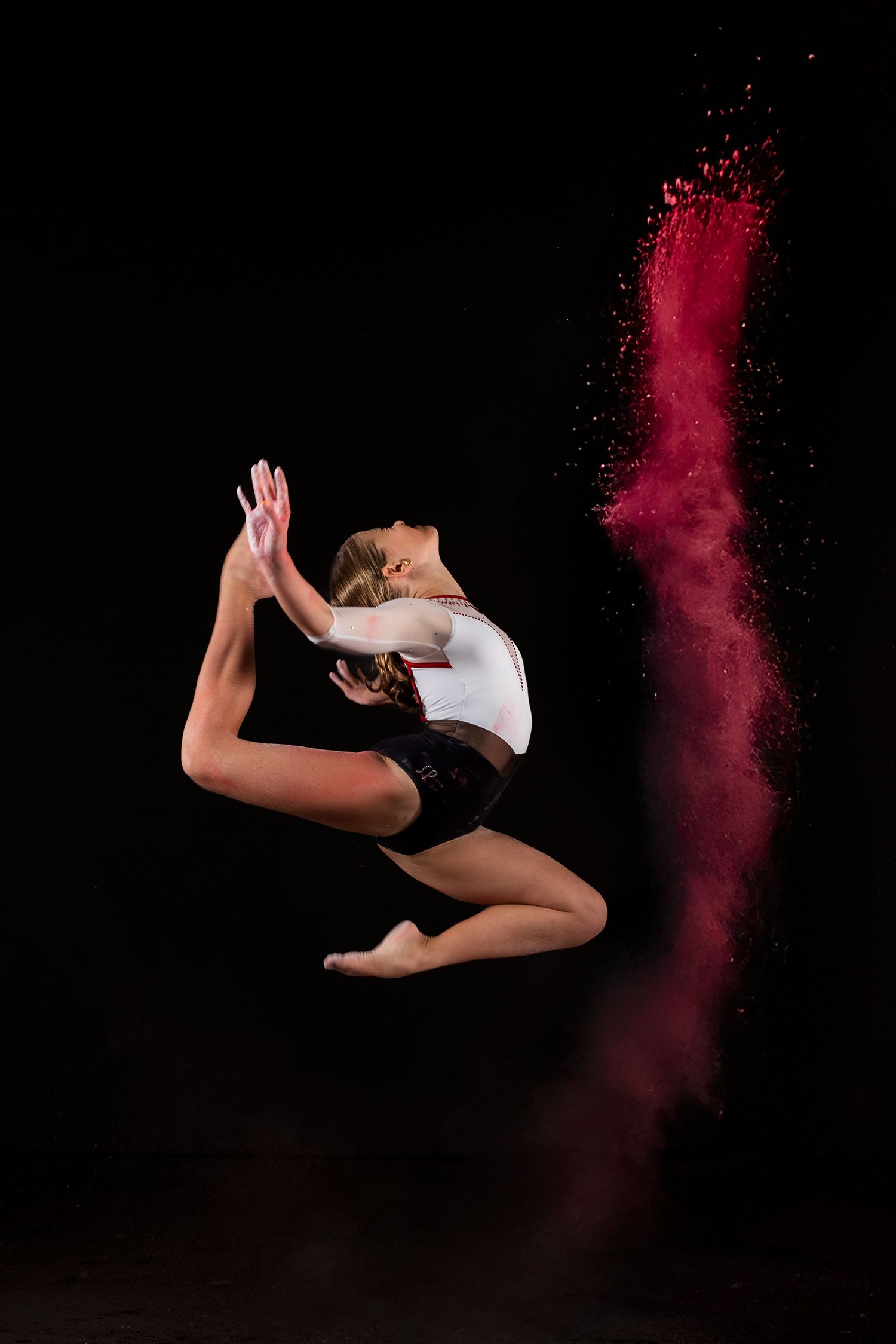Dynamic dancer in white top and black shorts leaps gracefully against dark background with red powder effect trailing behind.
