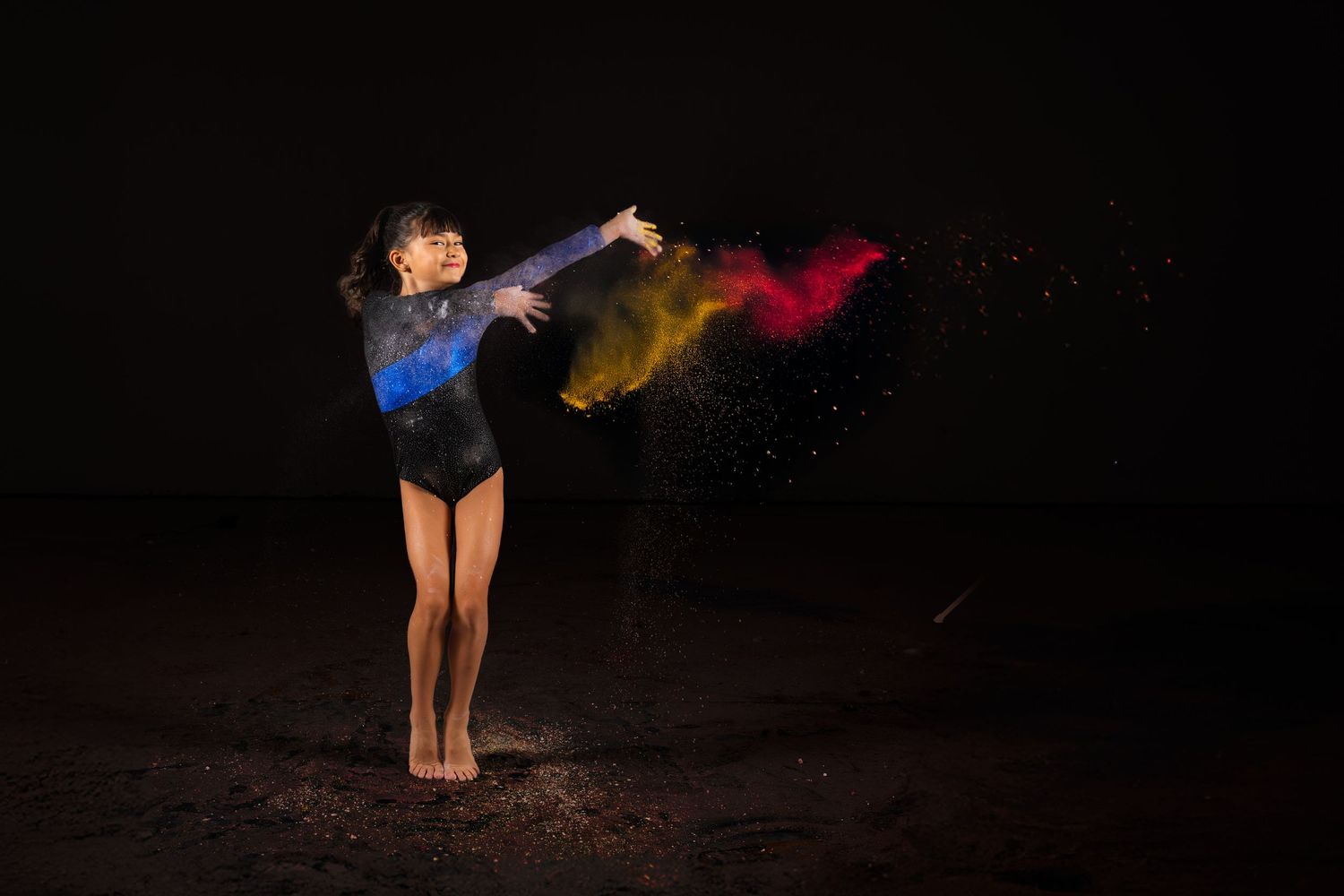 Dancer in blue leotard swirling colorful powder against dark background during dynamic performance shot.