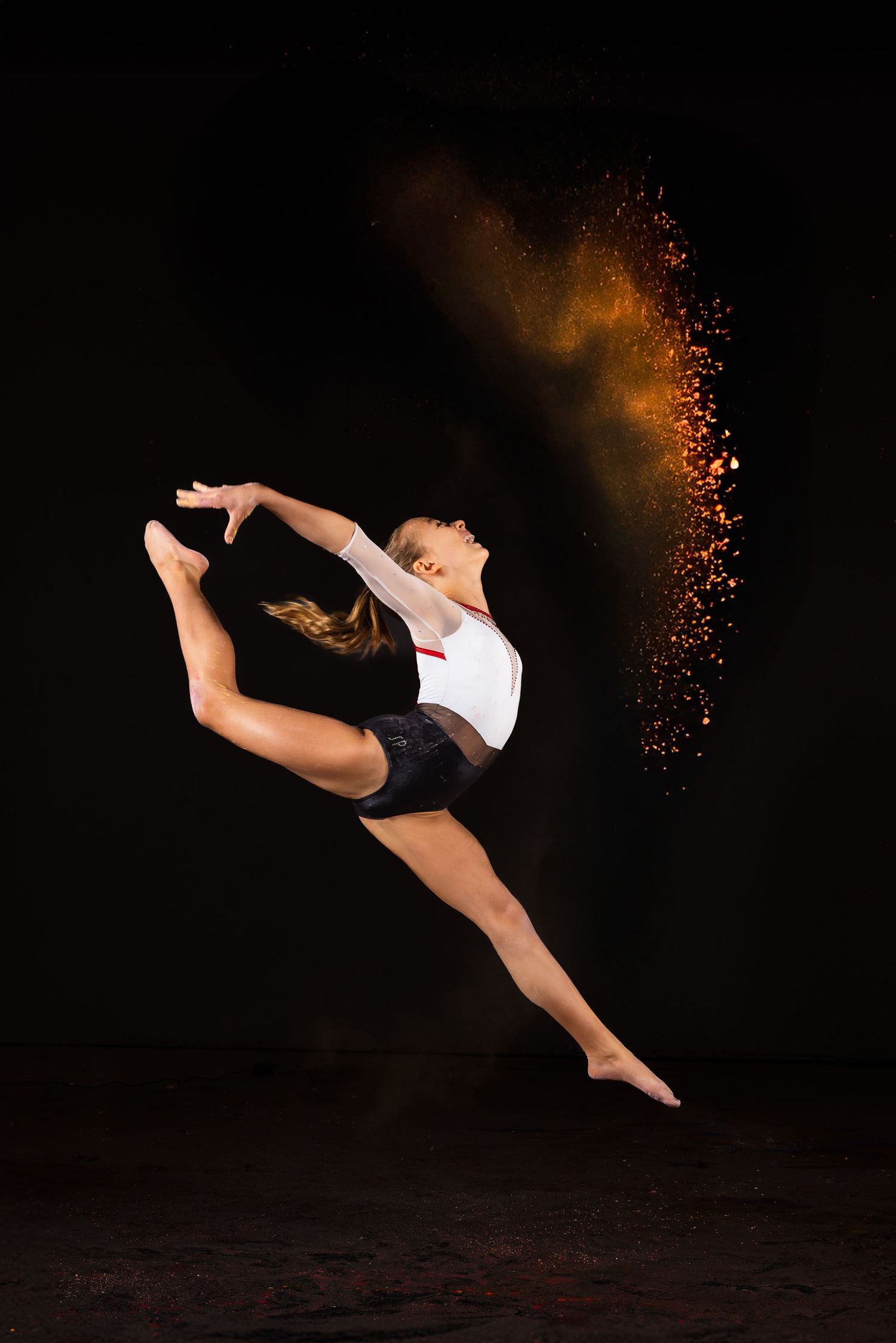 Graceful dancer in white and black performs a dramatic leap with a trail of glowing orange sparks against dark background.
