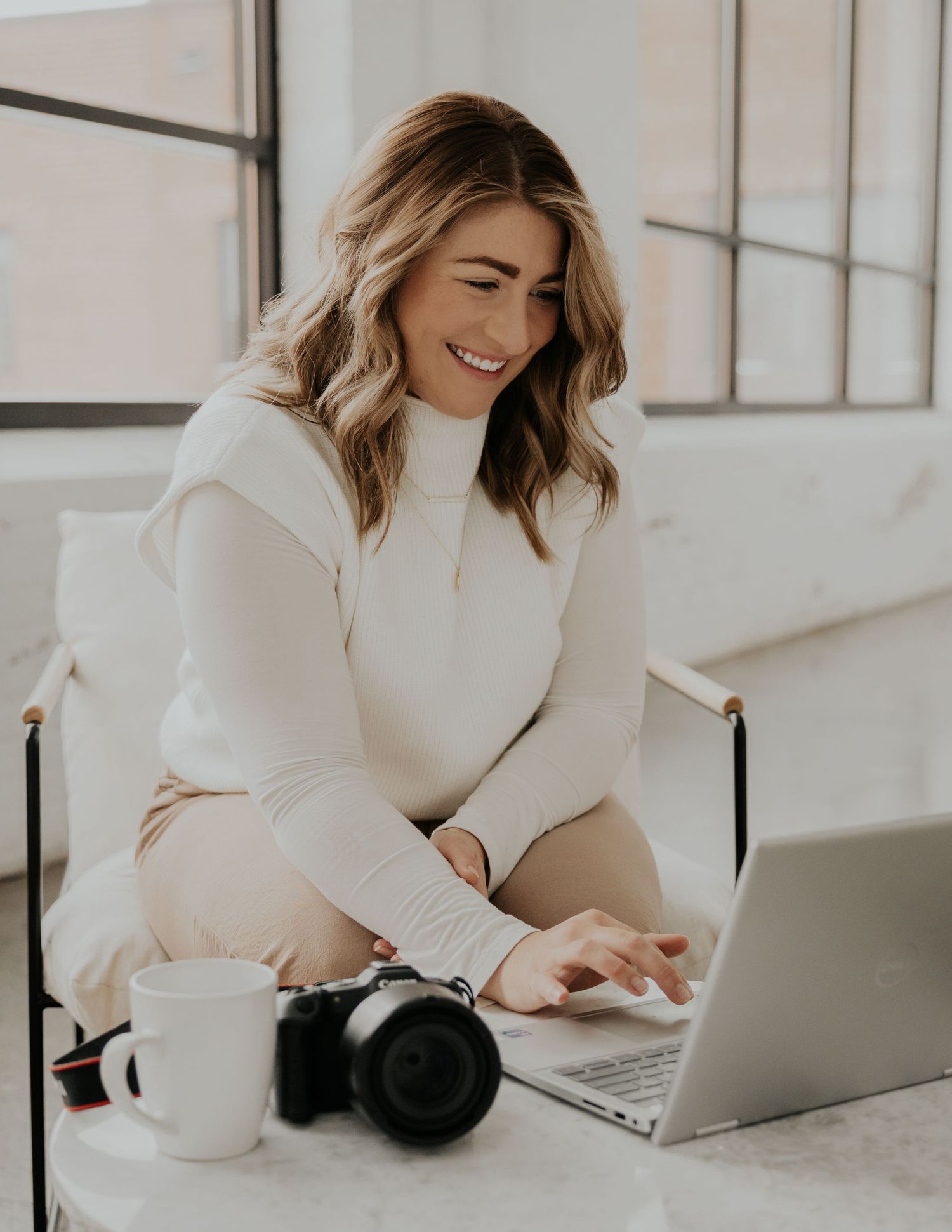 Person in white sweater works on laptop with camera on desk while smiling in bright minimalist setting.