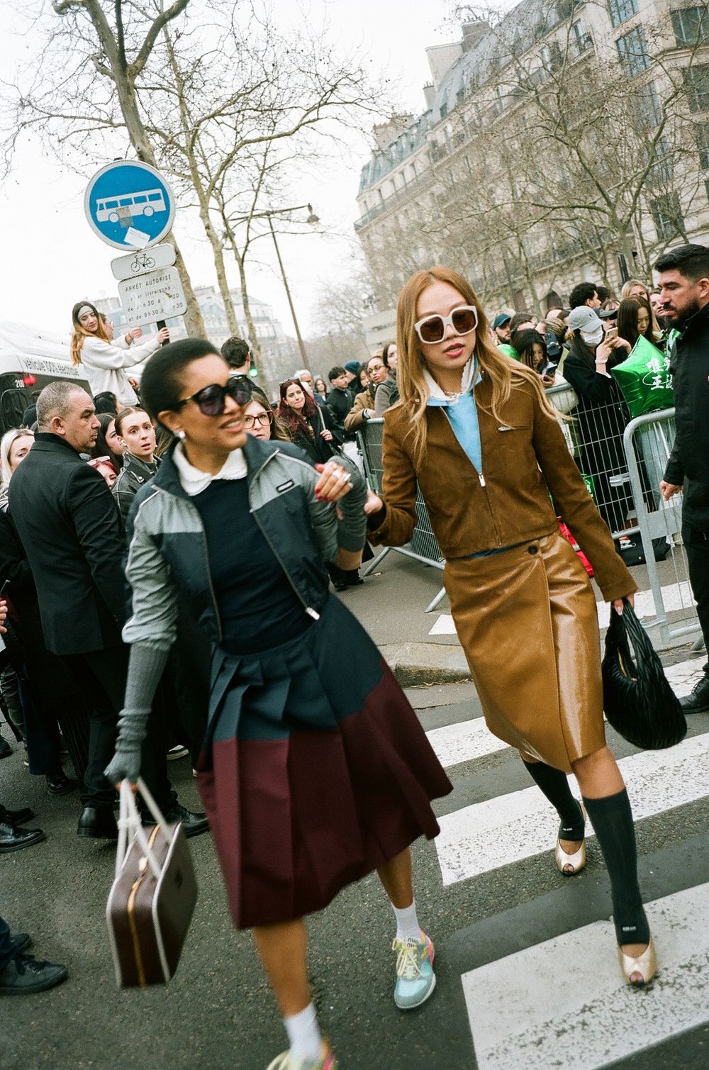 Two people in stylish coats walk across a busy Paris street during fashion week, with crowds visible in background.