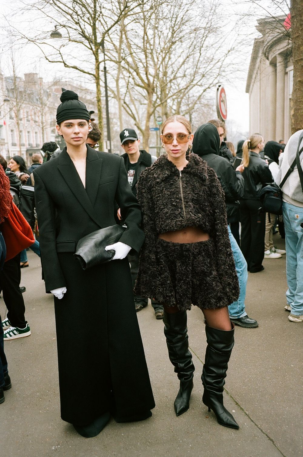Two fashionable people in dark winter outfits pose on a crowded street, one in long coat and one in cropped fur ensemble.
